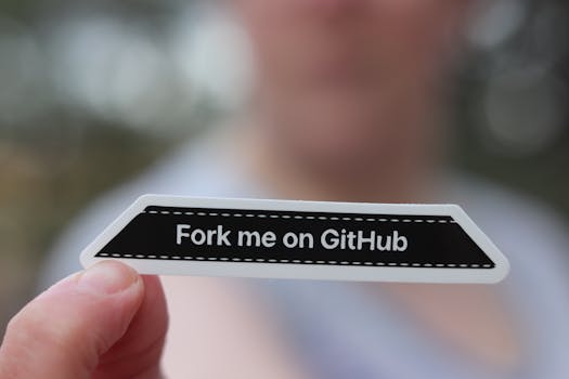 Close-up of a hand holding a 'Fork me on GitHub' sticker, blurred background.