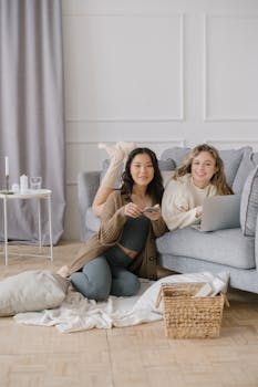 Two friends relaxing at home using a laptop and smartphone, showcasing comfort and connectivity indoors.