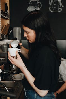A barista with long dark hair pouring milk into a coffee cup in a cozy Polish cafe setting.