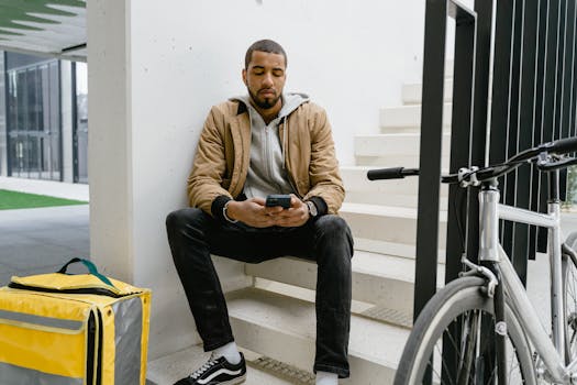 Young courier sits on stairs with phone, bike, and delivery bag, waiting in urban setting.